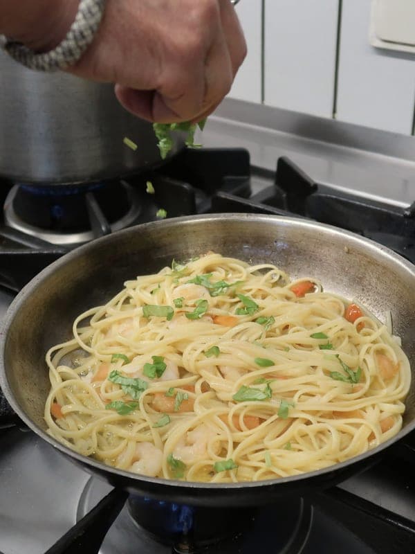 Eine Hand streut frisches Basilikum über eine Pfanne mit Linguine und Garnelen